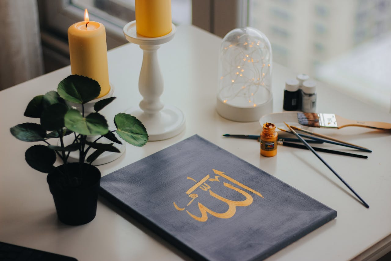A calming indoor scene showing Islamic calligraphy art, candlelight, plant, and painting tools.