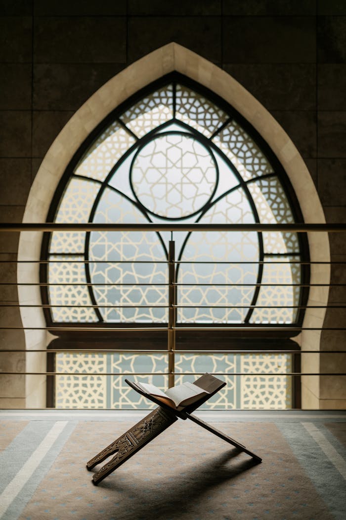 Open Quran on a wooden stand in front of an ornate arched window inside a mosque.